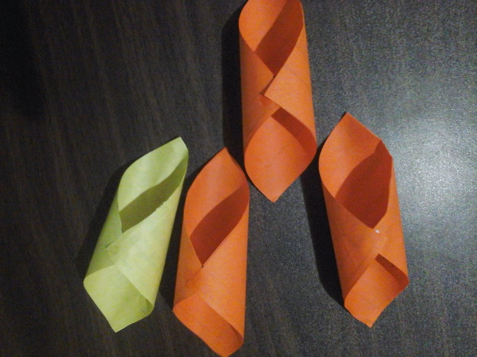 Four cone-shaped folded paper pieces, one light green and three orange, arranged on a dark textured surface